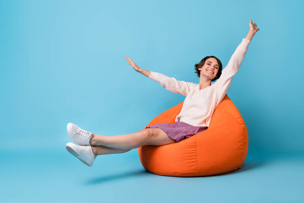 Photo portrait full body view of girl waking up stretching sitting in orange beanbag chair isolated on pastel blue colored background - Fotografie, Obrázek