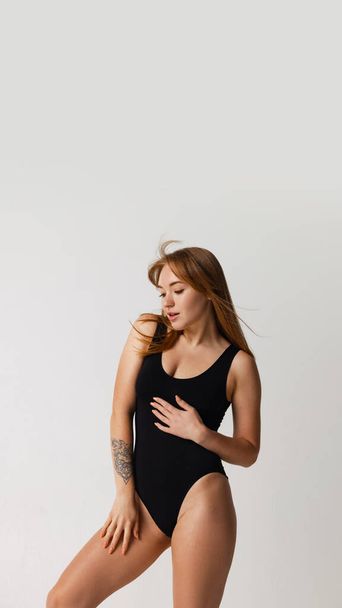 Portrait of young beautiful red-headed girl in black swimsuit posing isolated over gray studio background. Natural beauty concept. - Foto, imagen