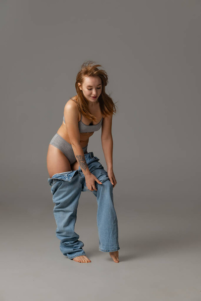 Young beautiful red-headed woman putting blue jeans jumpersuit on isolated over gray studio background. Weight Loss concept. - Foto, Imagem