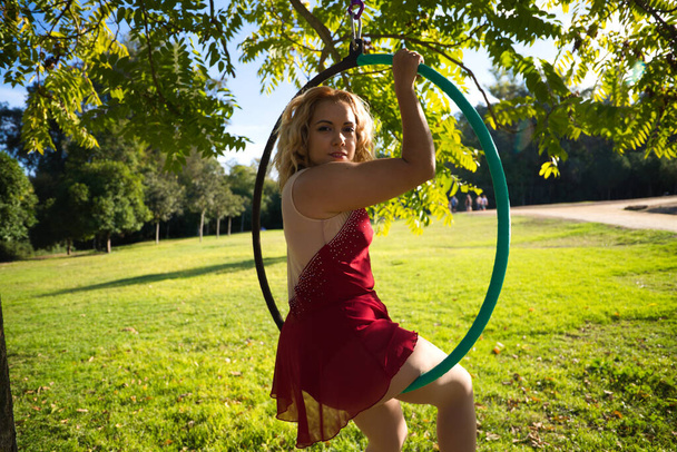 Blonde Frau und junge Turnerin Akrobatin, die Luftübungen auf dem Luftring im Freien im Park macht. Flexible Frau im roten Kostüm führt Posen von Zirkusartisten auf, die mit Hüften tanzen. - Foto, Bild