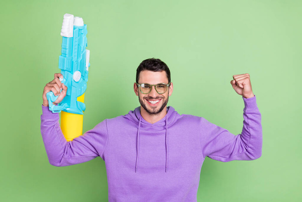 Photo of hooray millennial brunet guy hold watergun wear spectacles hoodie isolated on green color background - Fotoğraf, Görsel