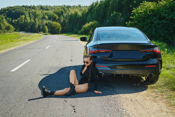 Sexy racer woman in bodysuit and rough boots sits on asphalt and leaning to her powerful racing car - 写真・画像