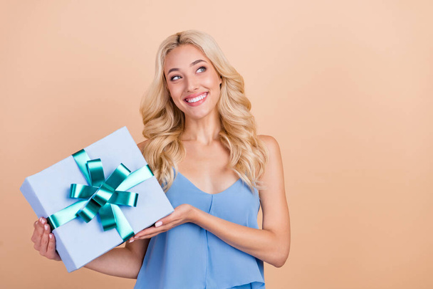 Photo of thoughtful sweet young lady wear blue singlet smiling holding gift box looking empty space isolated beige color background - Fotó, kép