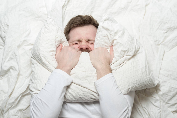 Man in white pajamas is stressed, has insomnia, cannot sleep, nervously covers mouth with a pillow and squeezes eyes shut. Top view.  - Photo, image