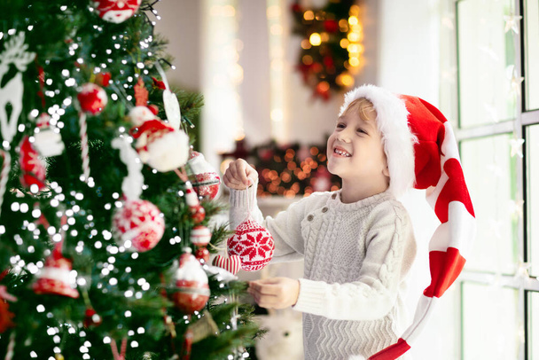 Παιδί στολίζει χριστουγεννιάτικο δέντρο στο σπίτι. Αγοράκι με πιτζάμες και χριστουγεννιάτικο στολίδι. Οικογένεια με παιδιά γιορτάζουν τις χειμερινές διακοπές. Τα παιδιά διακοσμούν το σαλόνι και το τζάκι για τα Χριστούγεννα. - Φωτογραφία, εικόνα