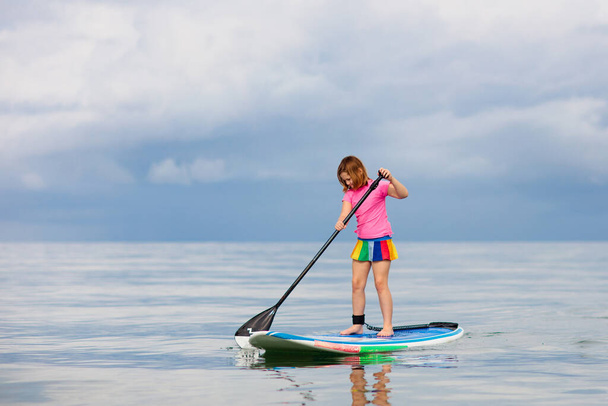 Child on stand up paddle board. Water fun and beach sport for kids. Healthy outdoor sports for summer vacation on tropical island. Holiday activity. Fit little girl training. Surfer exercising. - Foto, Imagem