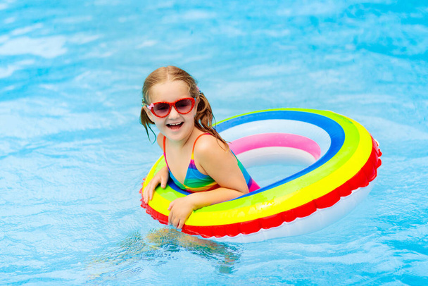 子供のプールおもちゃのリングに浮かんでいます。子供たちは泳ぐ。子供のためのカラフルな虹のフロート。トロピカルリゾートで家族の夏休みを楽しんでいる小さな女の子。ビーチや水のおもちゃ。太陽の保護. - 写真・画像