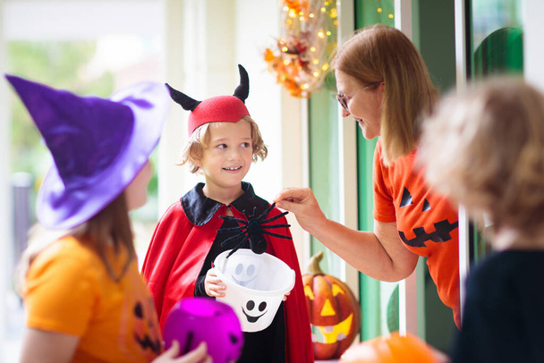 Dzieciaki zbierają słodycze w Halloween. Dziecko przy dekorowanych drzwiach domu. Chłopiec i dziewczyna w kostiumie czarownicy i wampira i kapeluszu z wiadrem cukierków i dyniową latarnią. Kobieta ze słodyczami.  - Zdjęcie, obraz