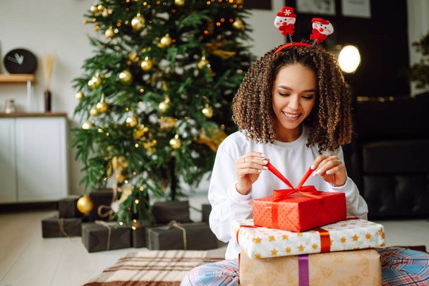 Afro American woman in pajamas and in santa claus hat with large gift box posing near Christmas tree. Winter holiday, vacation, relax and lifestyle concept. - Valokuva, kuva