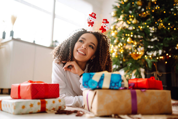 Afro American woman in pajamas and in santa claus hat with large gift box posing near Christmas tree. Winter holiday, vacation, relax and lifestyle concept. - Фото, зображення