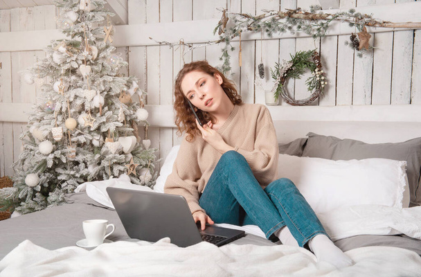 Caucasian brunette beautiful girl sits on bed with laptop, cup of coffee and christmas tree white - Foto, immagini