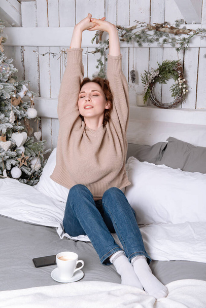 Valkoihoinen brunette kaunis tyttö sinisissä farkuissa ja beige pusero istuu sängyllä kupin kahvia valo joulukuusi kädet ylös kodikas - Valokuva, kuva