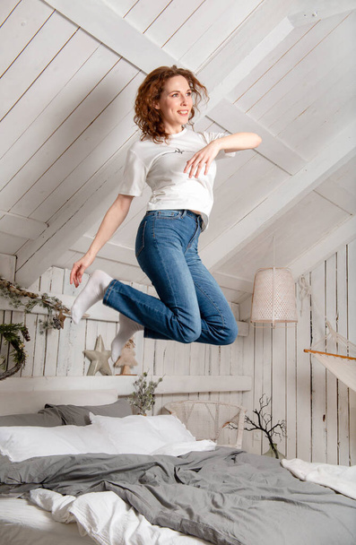 1 Avrupalı genç ve eğlenceli kadın, parlak bir odada, bir yatak tebessümünün üzerine atlıyor., - Fotoğraf, Görsel