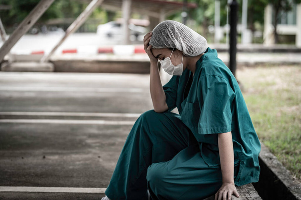 Tired depressed female asian scrub nurse wears face mask blue uniform sitting on hospital floor - Zdjęcie, obraz