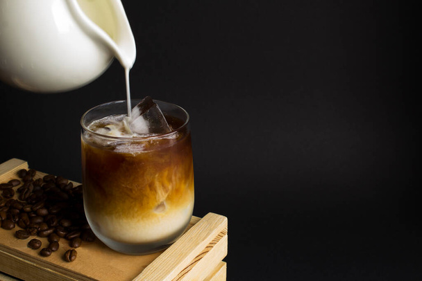Kalter Brühkaffee mit Milch und Eis im Trinkglas auf der Holzkiste auf schwarzem Hintergrund. Kopierraum. Nahaufnahme. - Foto, Bild