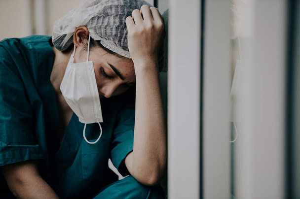 Väsynyt masentunut nainen aasialainen hangata sairaanhoitaja pukeutuu kasvot naamio sininen univormu istuu sairaalan lattialla - Valokuva, kuva