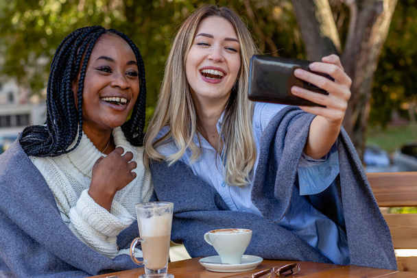 2人の魅力的な若い女性が一緒にコーヒーショップでコーヒーを楽しみ、テーブルに座ってスマートフォンで自撮りします。多様性と多民族概念. - 写真・画像