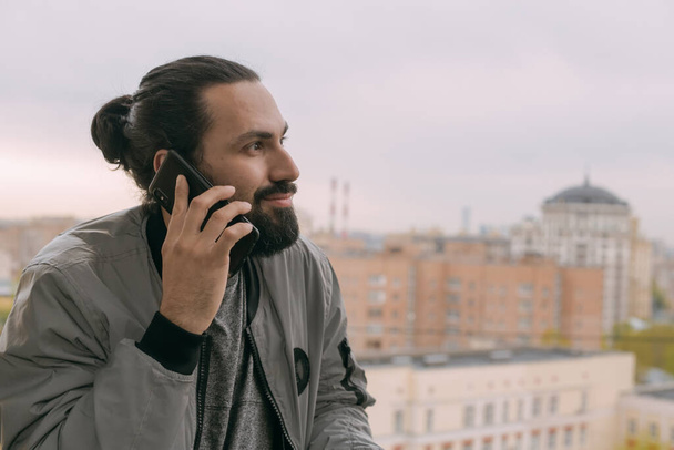 Egy fiatal, jóképű férfi portréja, telefonnal az erkélyen, kilátással a városra. Egy hosszú hajú fickó egy okostelefonon kommunikál a lakása vagy irodája erkélyén. - Fotó, kép