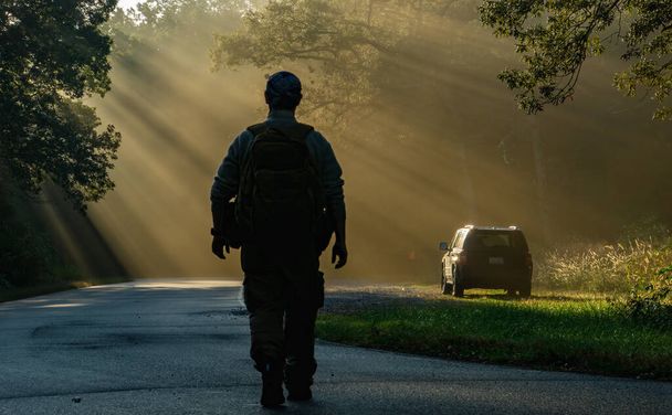 Mann unterwegs mit einem Rucksack auf dem Rücken. Sonnenstrahlen im Hintergrund mit einem am Straßenrand geparkten Fahrzeug, auf das er in einem Waldstück zugeht. - Foto, Bild