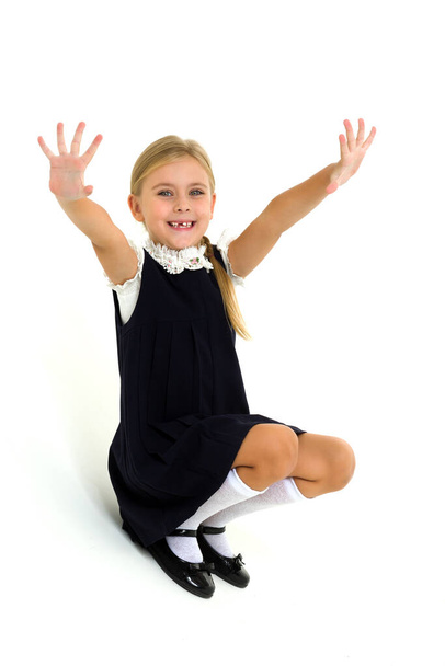 A cheerful blonde in a school uniform is sitting on the floor and waving her hands - Φωτογραφία, εικόνα