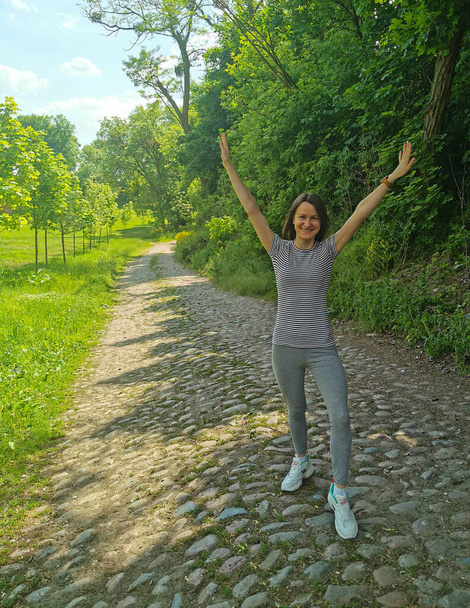 Usmívající se mladá dívka v parku na chodníku se zdviženýma rukama. Krásný slunný park se zelenými stromy - Fotografie, Obrázek