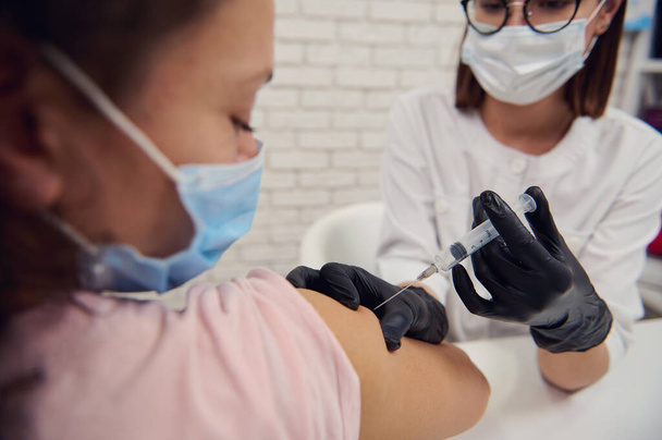 Siyah saçlı, koruyucu maskeli bir kadın Corona virüsü aşısıyla aşılanıyor. Bulaşıcı viral hastalıkların yayılması için gönüllü aşı kavramı. Nüfusun dokunulmazlığı - Fotoğraf, Görsel
