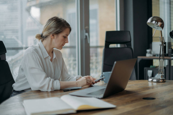 Молодая женщина с ноутбуком и телефоном за рабочим столом в современном офисе днем. Женщина в строгой офисной одежде днем работает за компьютером на рабочем месте - Фото, изображение