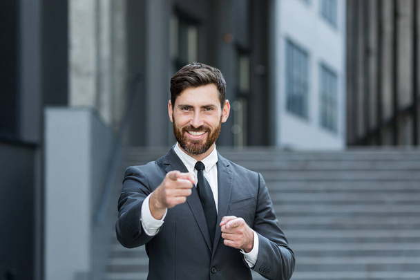 Portrait of young successful businessman looking at camera and smiling outdoors holding thumbs up rejoicing - Photo, Image