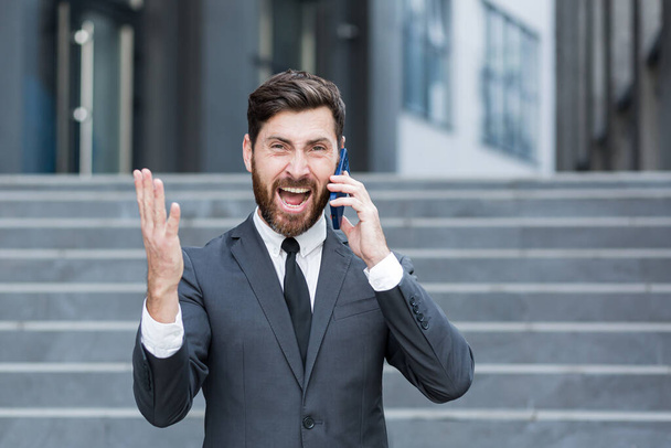 angry male businessman cry talking on the phone outside business man shouting on call outdoors. Nervous mad professional conversation on smartphone. Irritated employee scream speak at urban street - Φωτογραφία, εικόνα