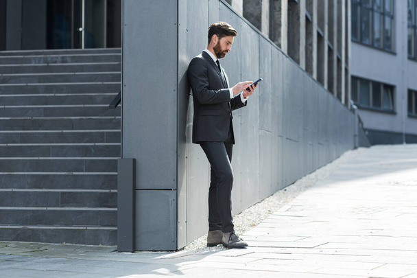 Caucasian bearded employee on urban background office building texting Messages, read good news mobile phone. Business man standing a formal suit messaging using Smartphone cellphone outdoors - Foto, Imagen