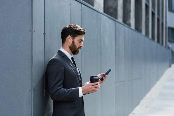 Caucasian bearded employee on urban background office building texting Messages, read good news mobile phone. Business man standing a formal suit messaging using Smartphone cellphone outdoors - Fotografie, Obrázek