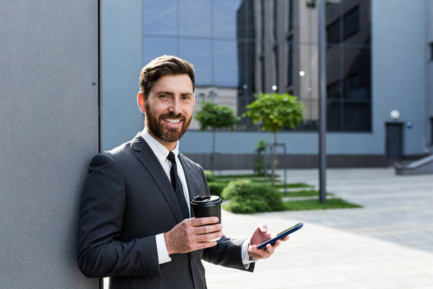 Successful businessman broker in a business suit talking on the phone near the office outside, Banker holding a cup of coffee during lunch break smiling - Fotoğraf, Görsel