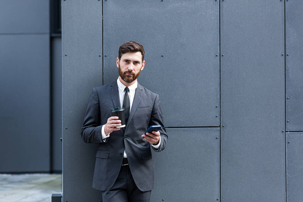 Bearded businessman in formal suit on break using mobile phone use smartphone. business man standing outside on modern urban city street background with coffee cup in downtown outdoors. copy space - Fotó, kép