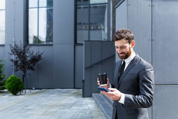 Caucasian bearded employee on urban background office building texting Messages, read good news mobile phone. Business man standing a formal suit messaging using Smartphone cellphone outdoors - Photo, Image
