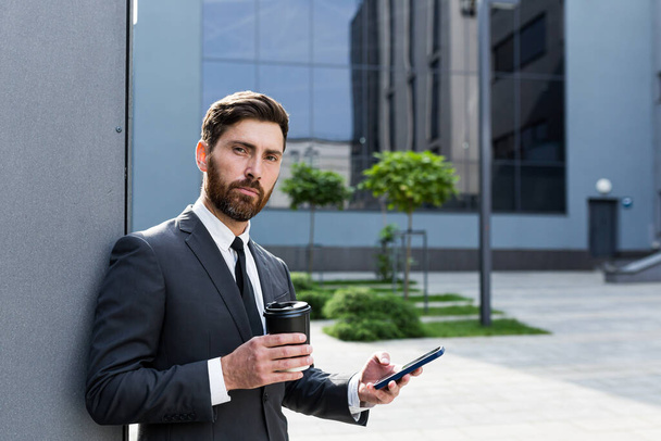 Successful businessman broker in a business suit talking on the phone near the office outside, Banker holding a cup of coffee during lunch break smiling - Foto, Imagem
