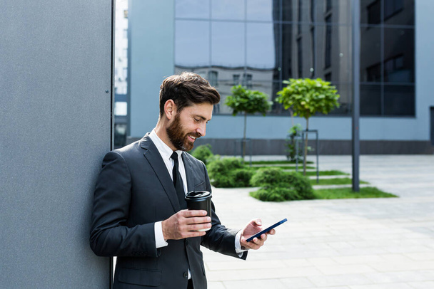 Bearded businessman in formal suit on break using mobile phone use smartphone. business man standing outside on modern urban city street background with coffee cup in downtown outdoors. copy space - Valokuva, kuva