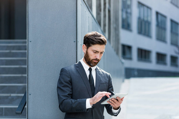 stylish bearded businessman in formal business suit standing working with tablet in hands on background modern office building outside. Man using smartphone or uses mobile phone outdoors city street - Foto, imagen