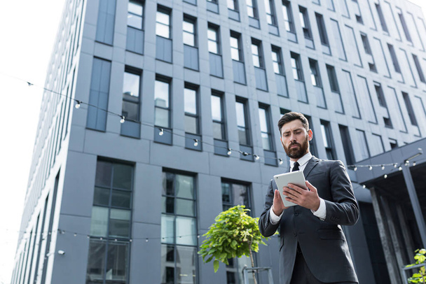 stylish bearded businessman in formal business suit standing working with tablet in hands on background modern office building outside. Man using smartphone or uses mobile phone outdoors city street - Foto, Imagem
