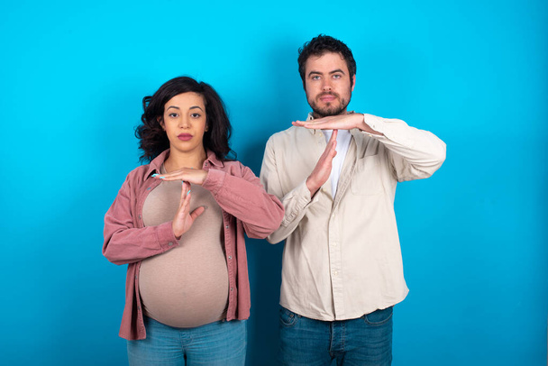 young couple expecting a baby standing against blue background being upset showing a timeout gesture, needs stop, asks time for rest after hard work, demonstrates break hand sign - Photo, Image