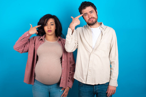 Unhappy young couple expecting a baby standing against blue background makes suicide gesture and imitates gun with hand, curves lips keeps two fingers on temple, shoots, being tired of everything, - Photo, Image