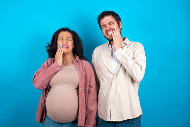 jeune couple s'attendant à un bébé debout sur fond bleu touchant la bouche avec la main avec une expression douloureuse en raison de maux de dents ou de maladies dentaires sur les dents. - Photo, image