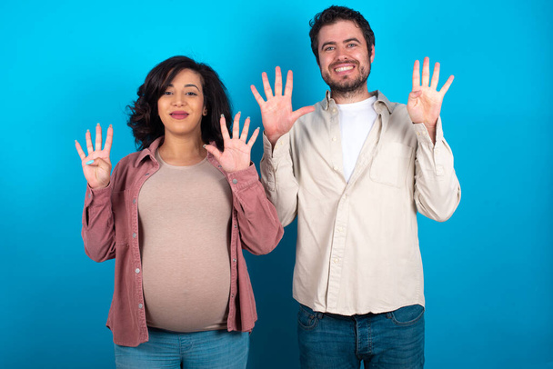 jeune couple attendant un bébé debout sur fond bleu montrant et pointant vers le haut avec les doigts numéro neuf tout en souriant confiant et heureux. - Photo, image