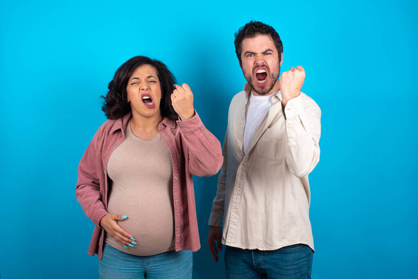 young couple expecting a baby standing against blue background angry and mad raising fist frustrated and furious while shouting with anger. Rage and aggressive concept. - Photo, Image