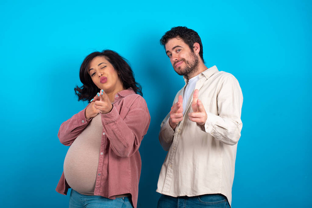 Joyful young couple expecting a baby standing against blue background wink and points index fingers at camera, chooses someone, makes finger gun pistol. - Photo, Image