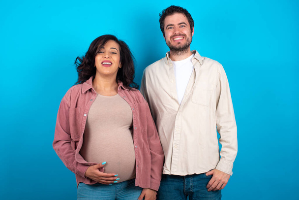 νεαρό ζευγάρι περιμένει ένα μωρό στέκεται πάνω σε μπλε φόντο με ένα χαρούμενο και δροσερό χαμόγελο στο πρόσωπο. Τυχερός άνθρωπος. - Φωτογραφία, εικόνα