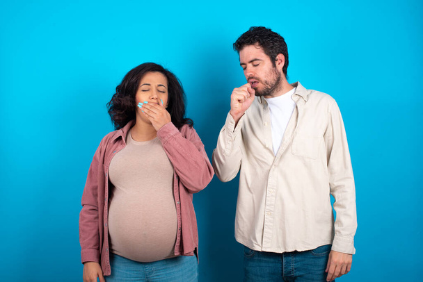 jeune couple s'attendant à un bébé debout sur fond bleu se sentant mal à l'aise et toussant comme symptôme de rhume ou de bronchite. Concept de soins de santé. - Photo, image