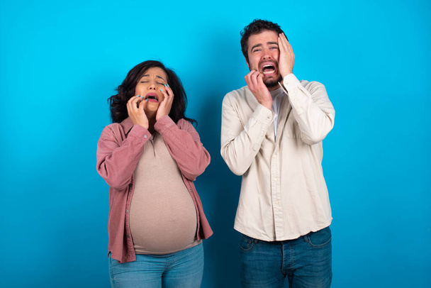 Doleful desperate crying young couple expecting a baby standing against blue background , looks stressfully, frowns face, feels lonely and anxious - Photo, Image
