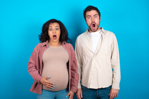 Oh mon Dieu ! Surpris jeune couple s'attendant à un bébé debout sur fond bleu regarde la caméra avec expression choquée s'exclame avec inattendu, - Photo, image