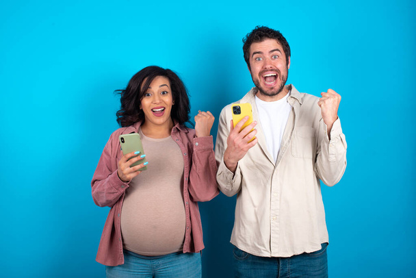Positive young couple expecting a baby standing against blue background holds modern cell phone connected to headphones, clenches fist from good emotions, exclaims with joy, - Photo, Image