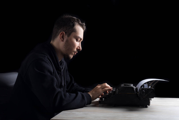 Nuori komea mies mustassa huppari kirjoittaa kirjoituskoneen, mustalla taustalla hillitty - Valokuva, kuva